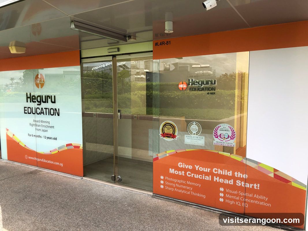 A view of the glass storefront entrance for Heguru Education. The facade features large orange and white vinyl decals on the glass panels. The left panel displays the company logo, the tagline 'Award-Winning Right Brain Enrichment From Japan', and the target age group 'For 6 months - 12 years old'. The right panel lists developmental benefits such as 'Photographic Memory' and 'High IQ, EQ', along with several award stickers on the glass. The entrance consists of automatic sliding glass doors.