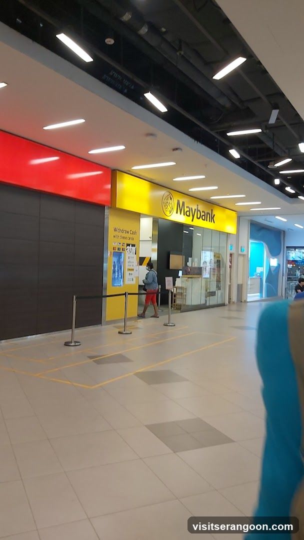 A view of a Maybank branch located inside a shopping mall or commercial building. The storefront features a prominent yellow sign with the Maybank logo and name. The entrance is open, revealing glass doors and interior counters. A person is standing near the entrance, and stanchions are set up on the tiled floor.