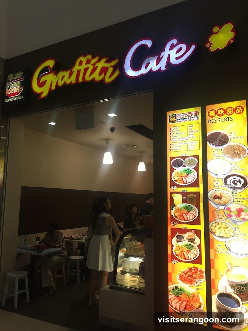 A brightly lit storefront with a large illuminated sign reading 'Graffiti Cafe' in stylized yellow and white lettering. To the right of the entrance, two vertical illuminated menu boards display various food and dessert options with images and prices. Inside, customers are seated at tables, and a glass display case is visible near the entrance.