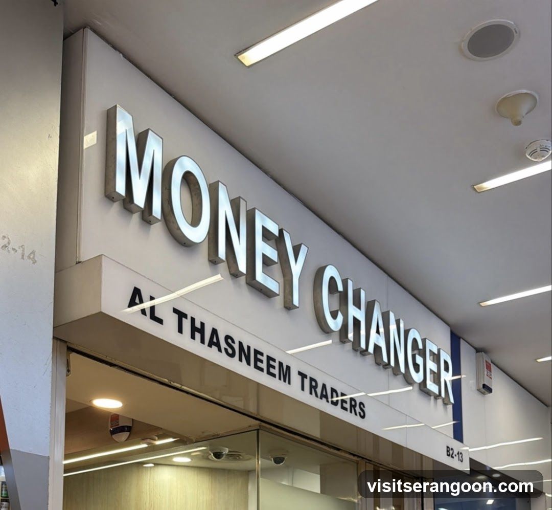A photo of a storefront with a large white sign reading 'MONEY CHANGER' in silver 3D letters. Below the main sign, smaller text reads 'AL THASNEEM TRADERS'. The shop number 'B2-13' is visible on the right side of the sign. The entrance features glass doors and interior lighting is visible.