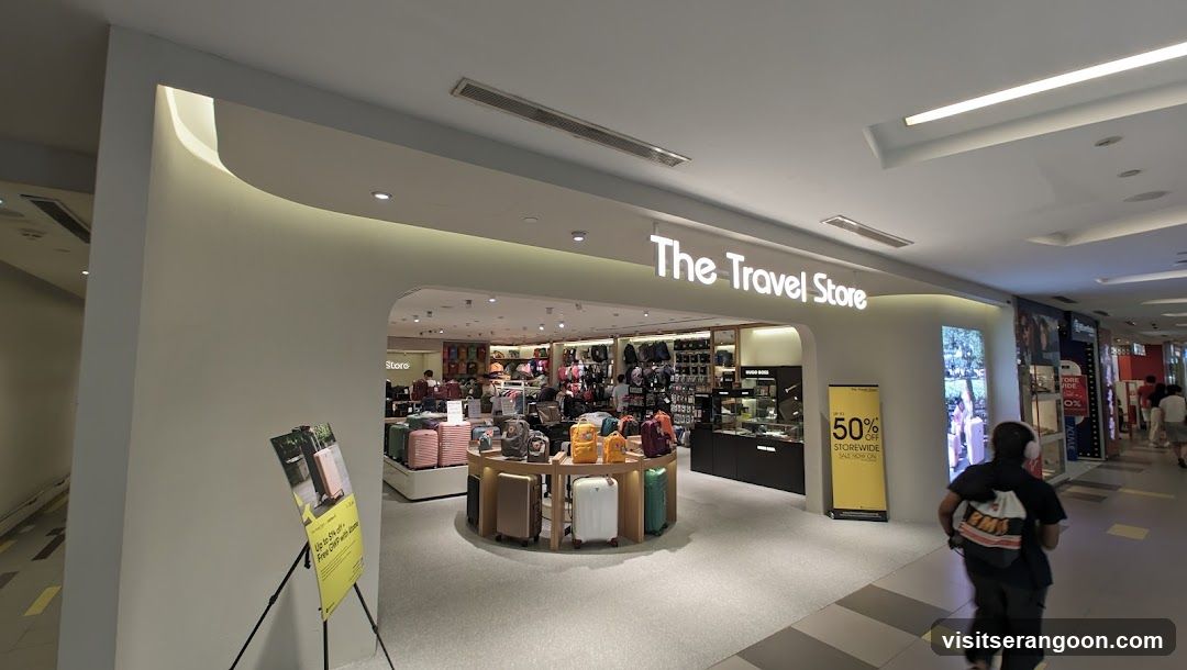 A brightly lit storefront with the illuminated sign 'The Travel Store' above the entrance. The interior displays various luggage and bags on shelves and a central circular display stand. A yellow promotional sign advertising '50% OFF STOREWIDE' stands near the entrance, and a person is walking past the store in the foreground.