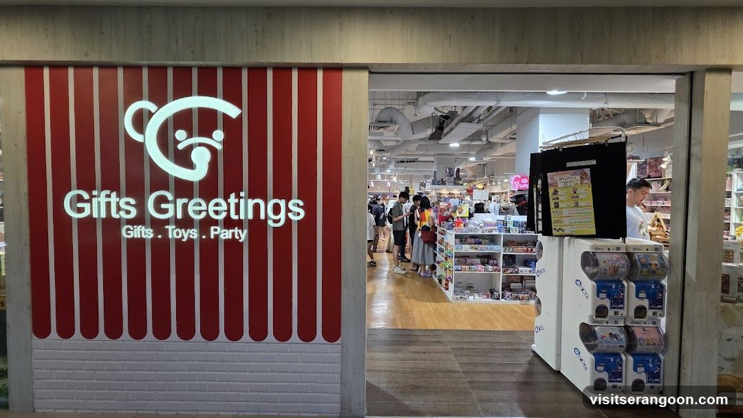 A view of a retail store entrance featuring a large red and white striped facade on the left. The wall displays a white logo of a bear head and the text 'Gifts Greetings' with the subtitle 'Gifts . Toys . Party'. The open doorway reveals the store's interior with wooden flooring, white shelving units stocked with merchandise, and several customers browsing. To the right of the entrance, there are white vending machines for capsule toys.