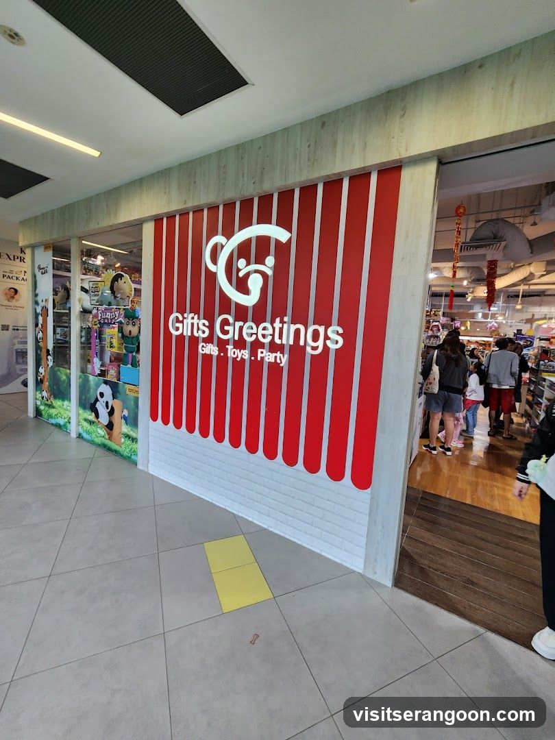 A view of a retail store entrance featuring a prominent facade with vertical red and white stripes. The store name 'Gifts Greetings' is displayed in large white letters along with the text 'Gifts. Toys. Party'. To the left, a window display shows various toys, including a large panda figure. Inside the open entrance, customers can be seen browsing aisles under warm lighting.