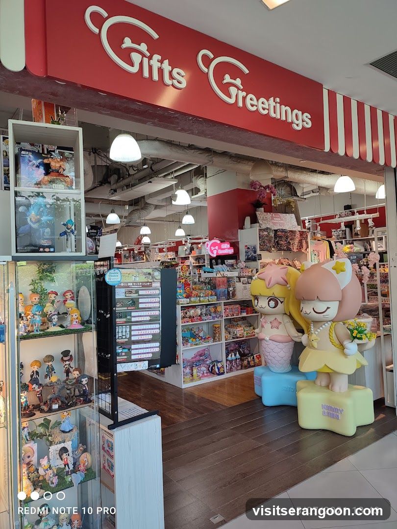 A view of the entrance to a retail store named 'Gifts Greetings'. The store features a large red sign with white lettering above the entrance. Inside, the shop is filled with shelves of merchandise, including boxed figures and collectibles. Two large, colorful statues of cartoon characters stand prominently near the entrance on the right side.