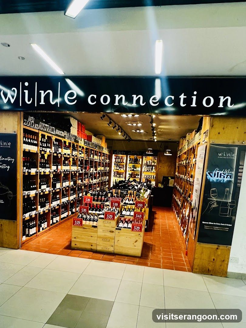 A brightly lit storefront for a business named 'wine connection'. The sign features white lowercase text on a black background. The entrance reveals a retail space with wooden shelving units fully stocked with wine bottles. In the foreground, wooden crates display bottles with red price tags, including prices like $39 and $49. The floor inside is tiled in terracotta, while the mall corridor outside has light-colored tiles.