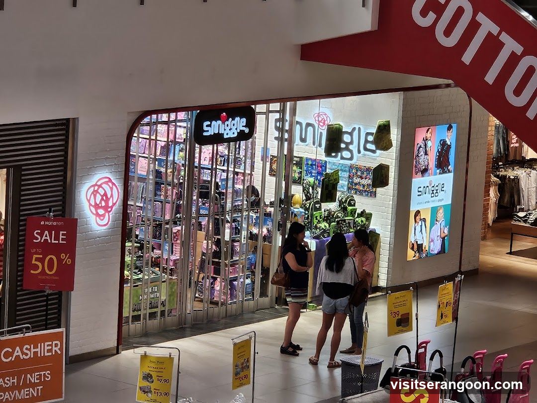 A brightly lit retail store entrance featuring the brand name 'smiggle' on a black sign above the door and in large white letters on the interior wall. The storefront has a white brick facade and glass doors. A red 'SALE up to 50%' sign is posted on the left, and yellow promotional signs stand on the floor near the entrance.