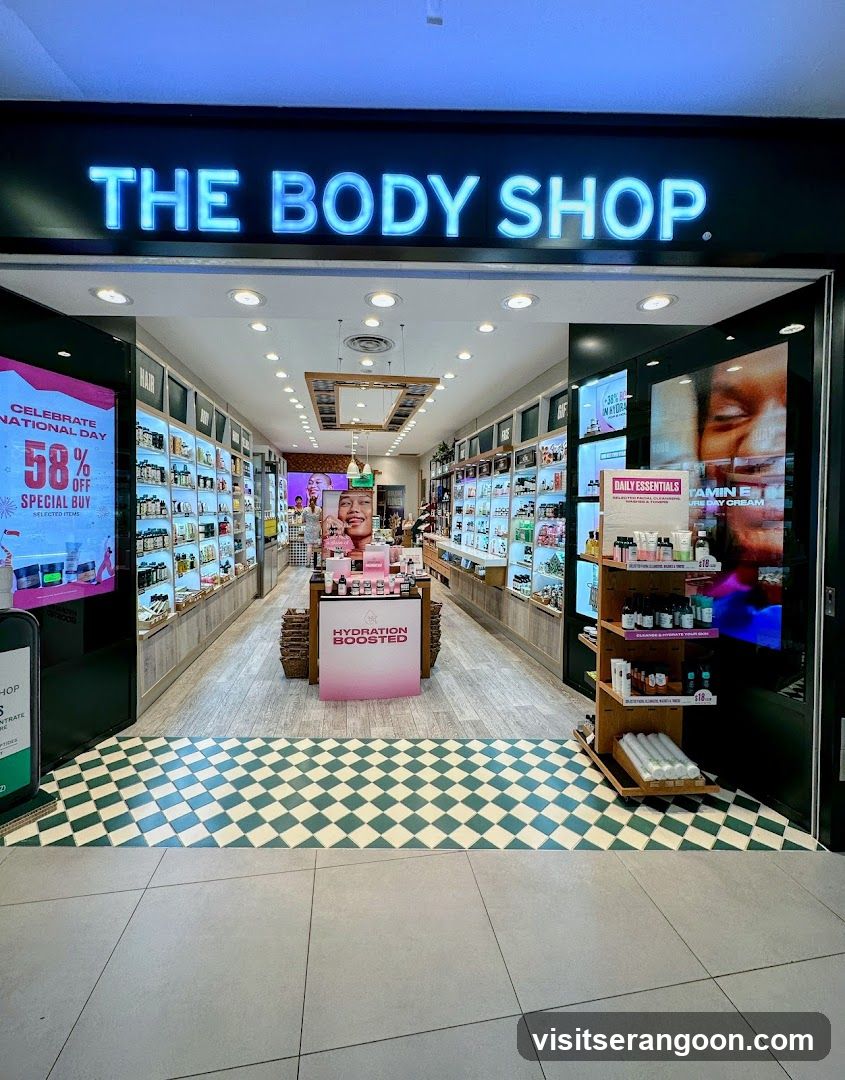 A brightly lit storefront for The Body Shop, featuring a large illuminated sign above the entrance. The interior is visible through open glass doors, displaying shelves of products, promotional posters, and a checkered floor pattern.