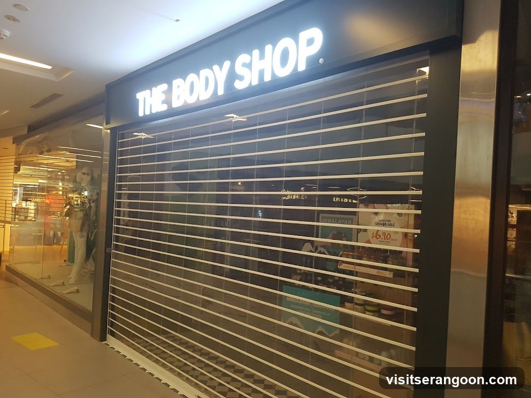A view of a closed retail store with a large, illuminated sign reading 'THE BODY SHOP' in white capital letters. The storefront is covered by a partially lowered metal rolling shutter with horizontal slats. Through the gaps in the shutter, shelves stocked with various products are visible inside the store. To the left, a glass window displays mannequins and other merchandise.