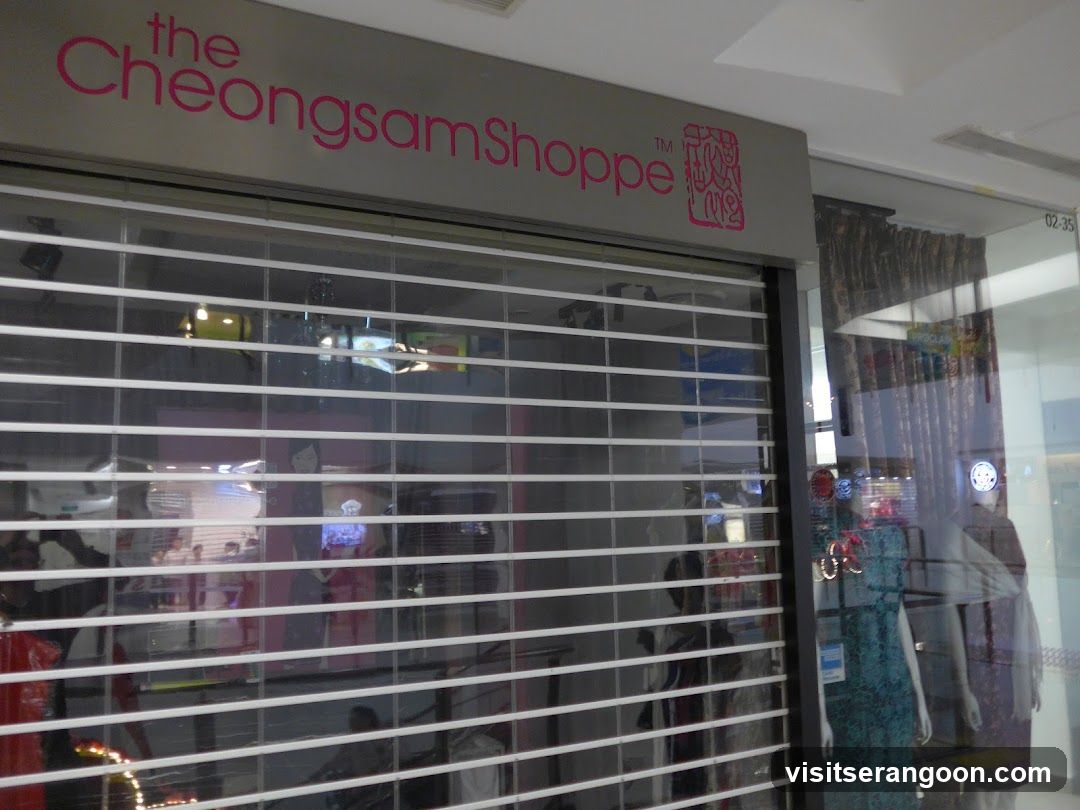 A storefront with a closed, white horizontal louvered security shutter. Above the shutter is a grey sign with the text 'the Cheongsam Shoppe' in pink lettering. To the right of the text is a pink logo resembling a traditional Chinese seal. Through the slats of the shutter, mannequins wearing dresses are visible inside the store.