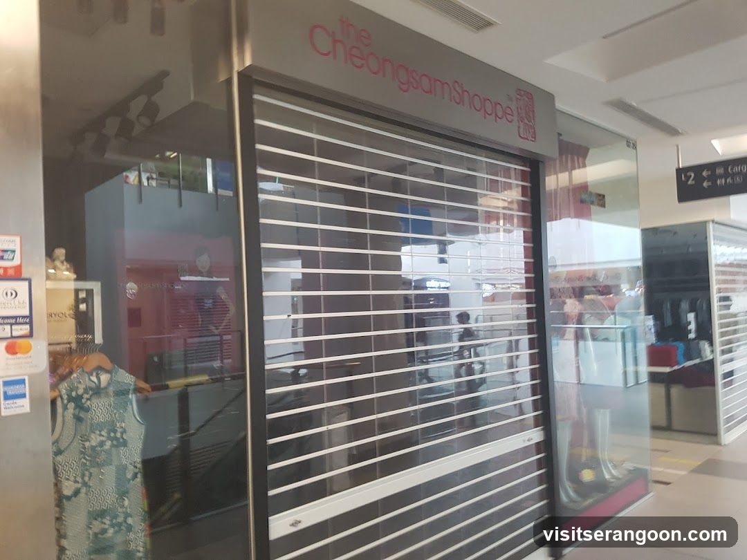 A closed storefront with a metal rolling shutter door. The sign above reads 'the Cheongsam Shoppe' in pink lettering. To the left, a glass window displays a patterned dress on a mannequin and payment stickers on the wall. To the right, a hallway leads to other stores, including one with a sign for '2'.