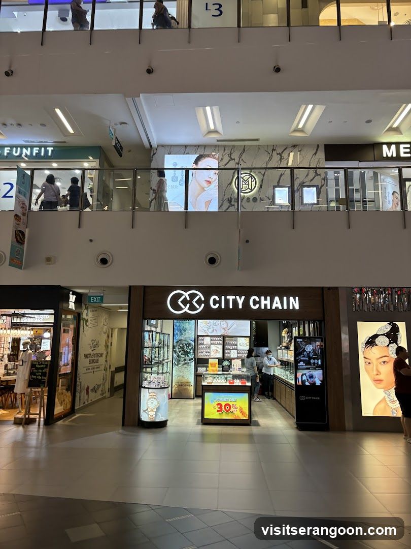 A wide shot of a City Chain jewelry store located on the ground floor of a multi-level shopping mall. The store features a dark brown facade with a prominent white illuminated sign reading 'CITY CHAIN' and its logo. Inside, glass display cases are visible, along with a promotional stand displaying a '30%' discount sign. To the left is another store with an 'EXIT' sign above it, and to the right is a large vertical advertisement featuring a model wearing jewelry. The upper levels show other shops, including 'FUNFIT' and 'ME', with people visible on the balconies.