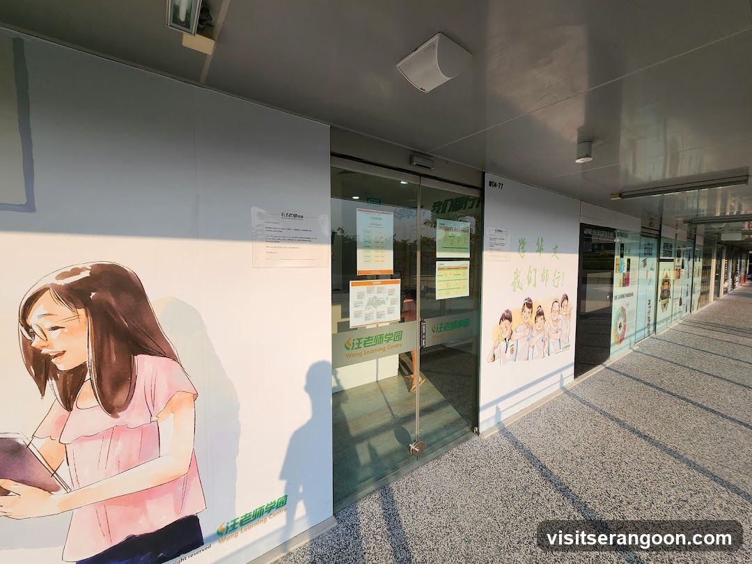 A view of a commercial hallway featuring a glass storefront entrance. The facade includes large wall graphics with illustrations of people and Chinese text, along with various posted notices and flyers on the glass doors and walls. A speaker is mounted on the ceiling above the entrance.