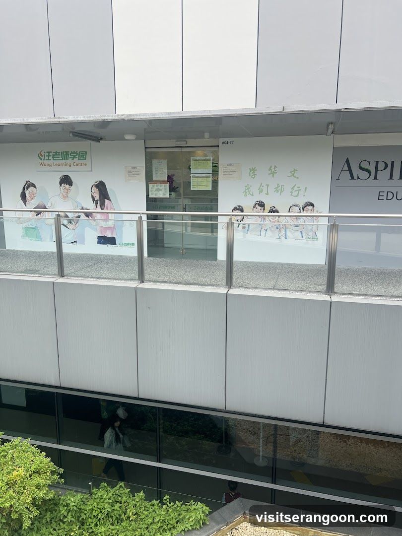 A view of a commercial storefront located on an upper level, viewed from a lower floor. The facade features large glass windows and doors with promotional posters. The left poster displays the text 'Wong Learning Centre' alongside illustrations of students. The center poster features Chinese text '学华文 我们都行!' (Learn Chinese, we can all do it!) with cartoon children. A partial sign for 'ASPIRE EDU' is visible on the right. A metal railing separates the viewer from the storefront.
