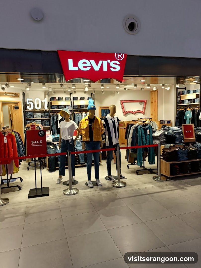 A view of a Levi's clothing store entrance inside a shopping mall. The store features a large red sign with the white 'Levi's' logo above the entrance. Inside, three mannequins are dressed in jeans and jackets, standing behind a red velvet rope barrier. Racks of clothing, including jeans and shirts, are visible on shelves and hangers. Sale signs are placed near the entrance.