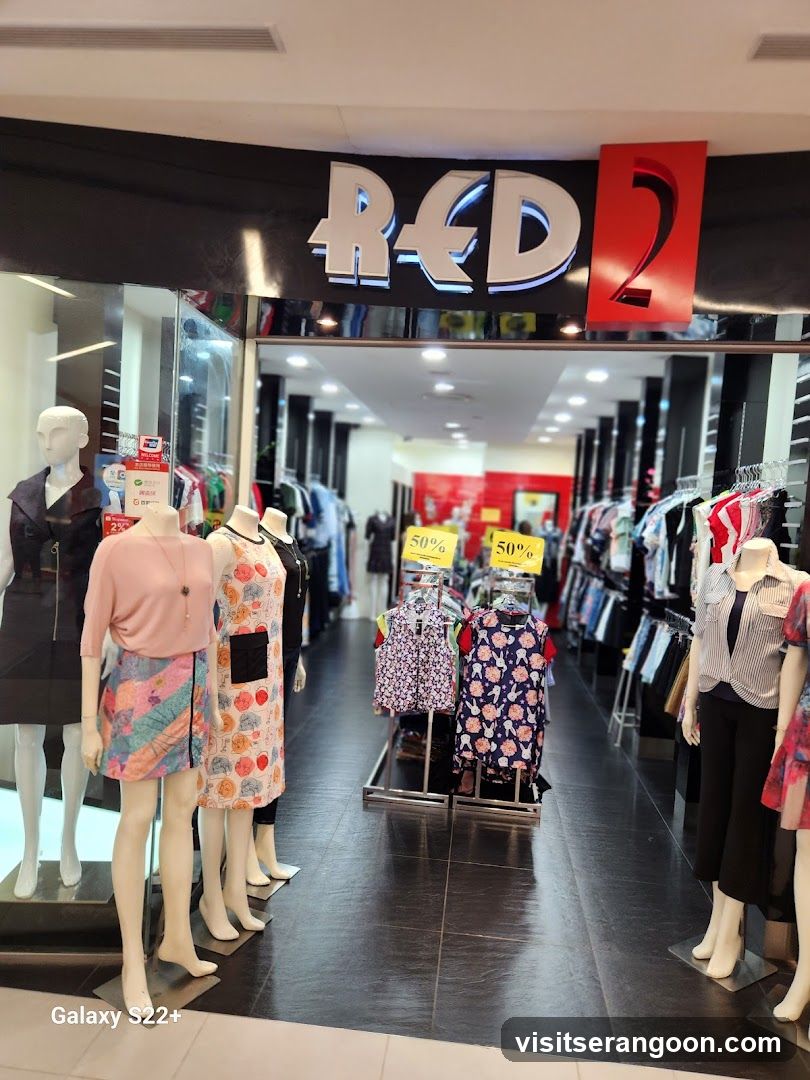 A view of the entrance to a clothing store named 'RED 2'. The store has a black facade with large, white 3D lettering for the name and a red square containing the number '2'. Inside, the store is brightly lit with rows of clothing racks and mannequins displaying various outfits. Yellow signs indicating '50%' discounts are visible on some displays.