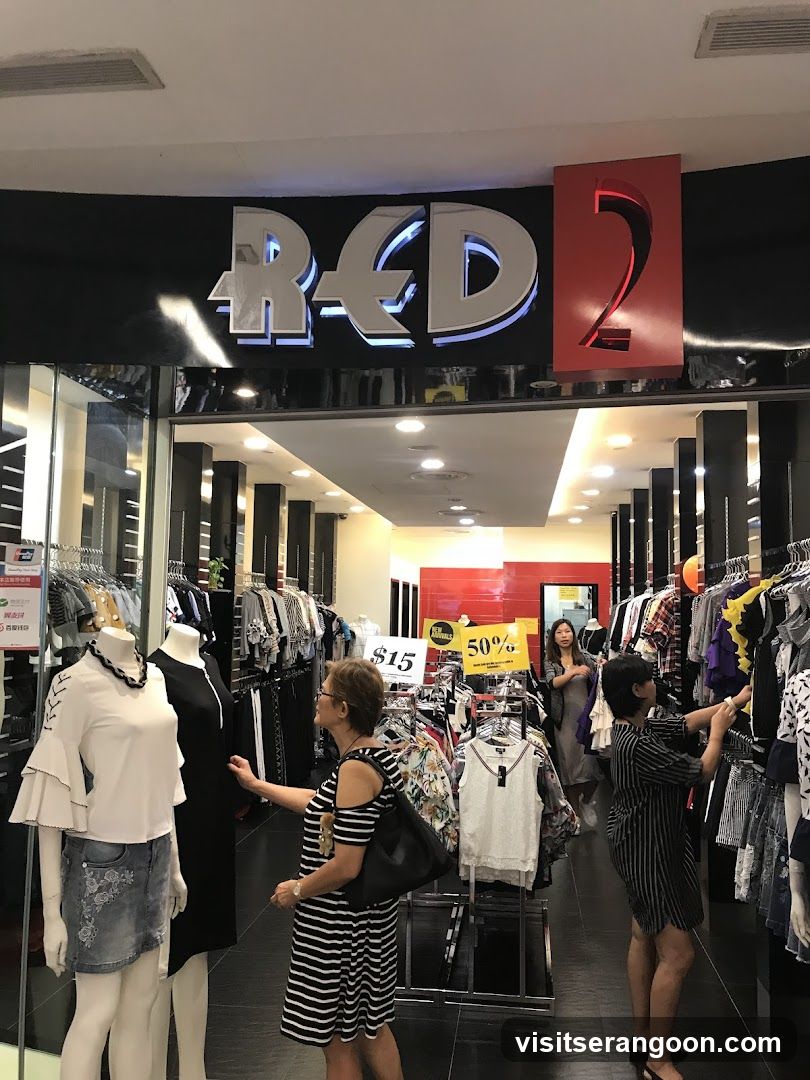 A view of the entrance to a clothing store named 'RED 2'. The store has a black facade with large, silver 3D lettering for the name and a red square containing the number '2'. Inside, the store is brightly lit with white ceiling lights. Several clothing racks are visible, filled with various garments. Three women are browsing the merchandise. A yellow sale sign indicating '50%' off is visible in the background.