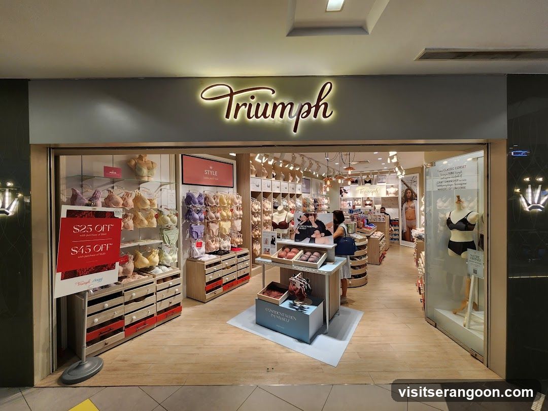 A brightly lit Triumph lingerie store entrance inside a shopping mall. The store features a large, illuminated 'Triumph' sign above the entrance. Inside, racks of bras and underwear are visible, along with promotional signage advertising discounts of $25 and $45 off. Mannequins display lingerie sets, and customers can be seen browsing the merchandise.