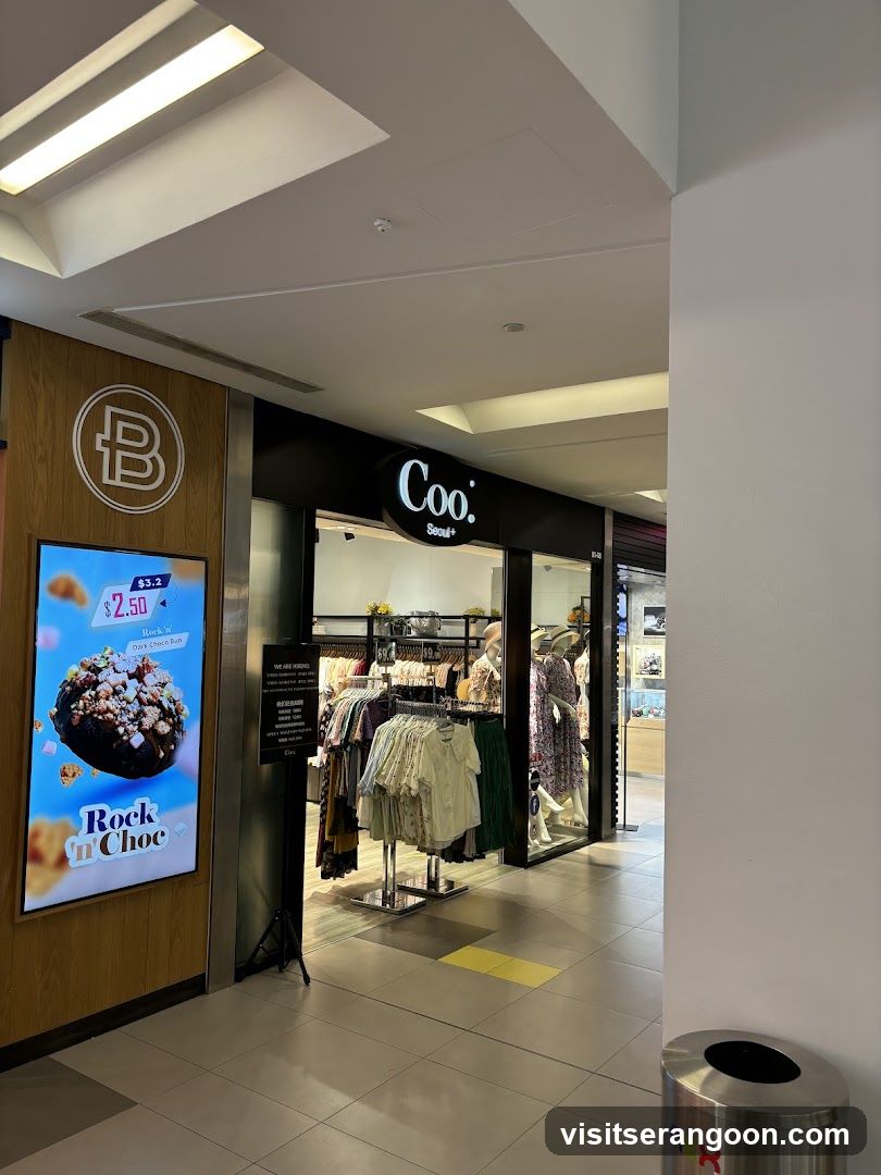 A view of a retail store entrance inside a shopping mall. The store has a black facade with the name 'Coo.' in white lettering. Inside, clothing racks and mannequins are visible. To the left, a digital advertisement displays a chocolate dessert.