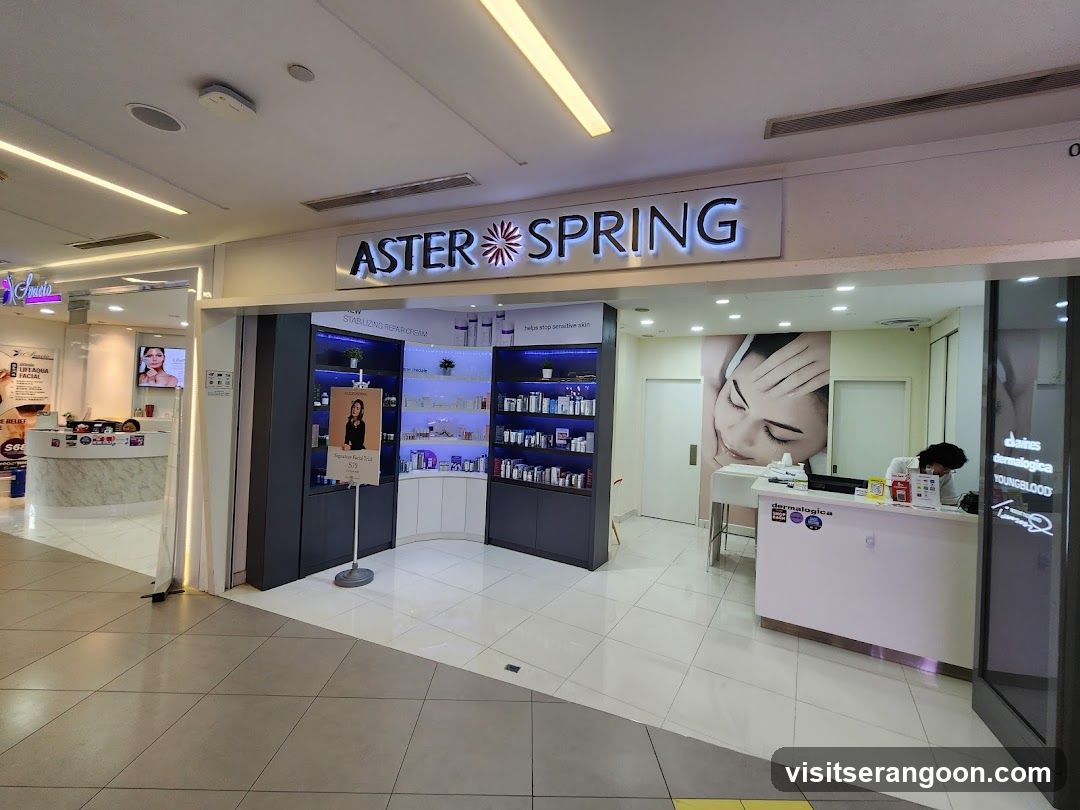 A brightly lit retail storefront with a large illuminated sign reading 'ASTER SPRING' above the entrance. The interior features white tiled floors, dark shelving units displaying various products, and a white reception desk on the right where a staff member is seated. A large poster of a woman's face is visible on the back wall.