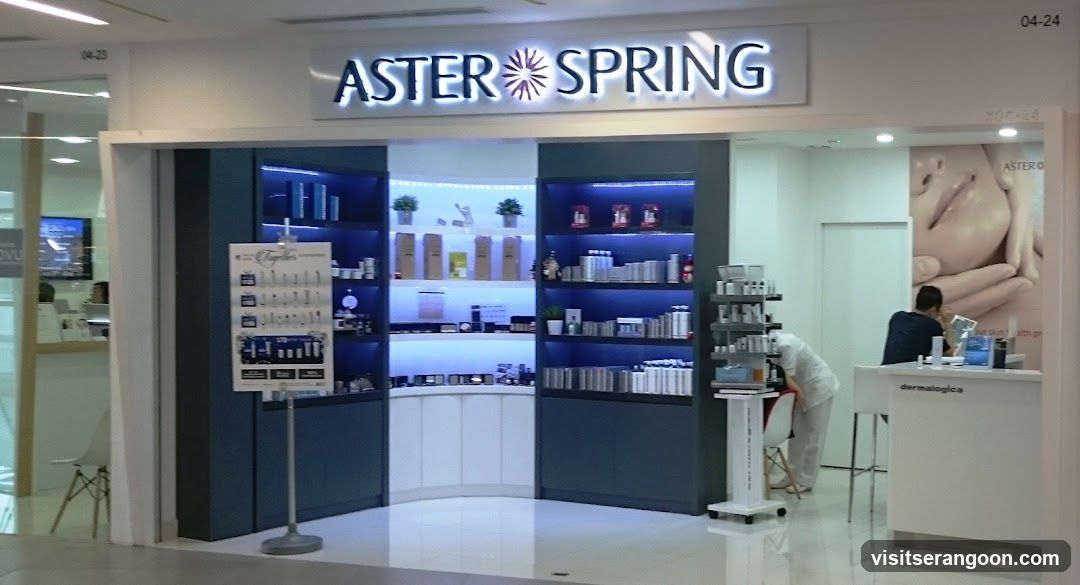 A brightly lit retail storefront with a large illuminated sign reading 'ASTER SPRING' above the entrance. The interior features dark blue shelving units displaying various skincare products, illuminated by blue accent lighting. A white reception desk is visible on the right with a staff member attending to a customer. A large poster of a face is mounted on the wall next to the desk.