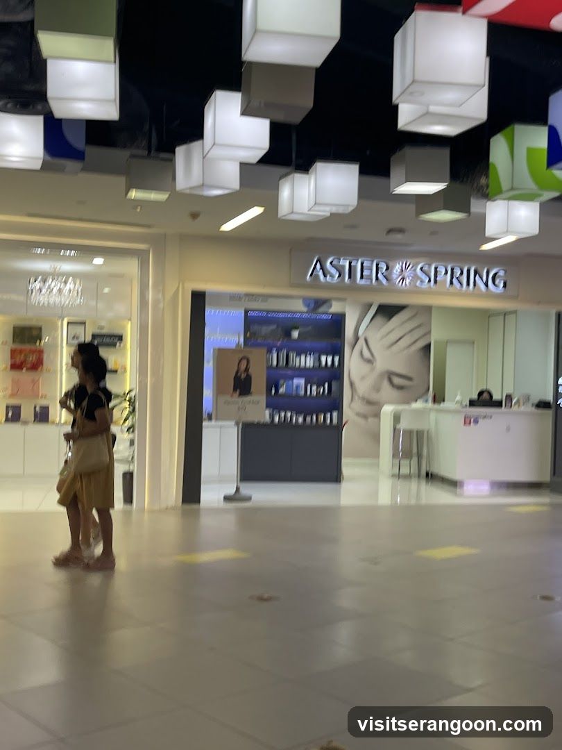 A view of the Aster Spring store entrance inside a shopping mall. The store features a white facade with the name 'ASTER SPRING' in illuminated lettering above the entrance. Inside, shelves of products are visible, along with a large wall graphic of a woman's face. A woman stands in the foreground near the store entrance.