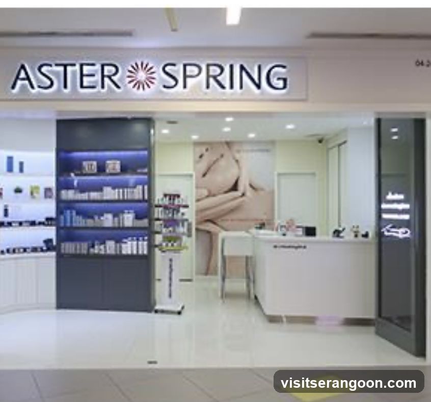 A brightly lit retail storefront with a large overhead sign reading 'ASTER SPRING' in dark lettering with a central flower logo. The entrance is open, revealing a clean interior with white tiled floors. To the left, a dark shelving unit displays rows of skincare products illuminated by blue backlighting. A white reception desk is visible on the right, and a large poster featuring a person's torso hangs on the back wall.
