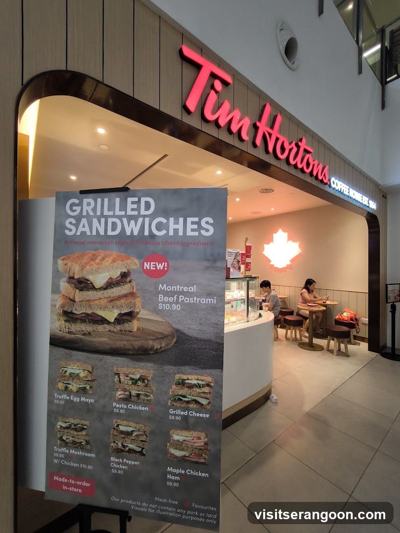 A view of a Tim Hortons storefront located inside a shopping mall. The store features a curved wooden facade with a large, illuminated red and blue sign reading 'Tim Hortons'. A large vertical poster board stands outside advertising 'GRILLED SANDWICHES' with images and prices for items like Montreal Beef Pastrami, Truffle Egg Mayo, and Pesto Chicken. Inside, customers are seated at tables near a counter.