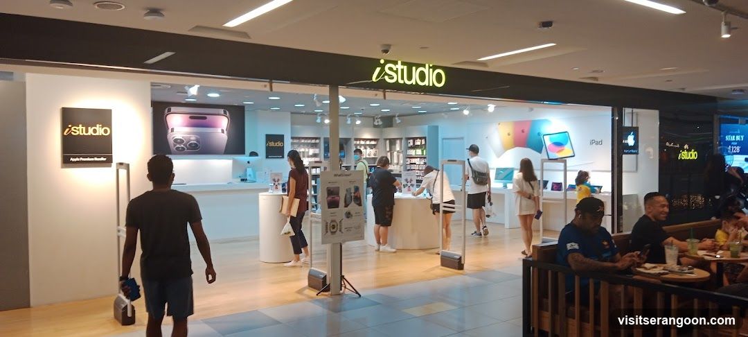 A wide-angle view of the iStudio electronics store entrance located within a shopping mall. The storefront features a large open glass facade with a prominent illuminated sign reading 'iStudio' in yellow lettering above the entrance. A smaller black sign with the same logo is mounted on the left wall. Inside, customers are browsing products and interacting with staff, while others sit at tables in the foreground.