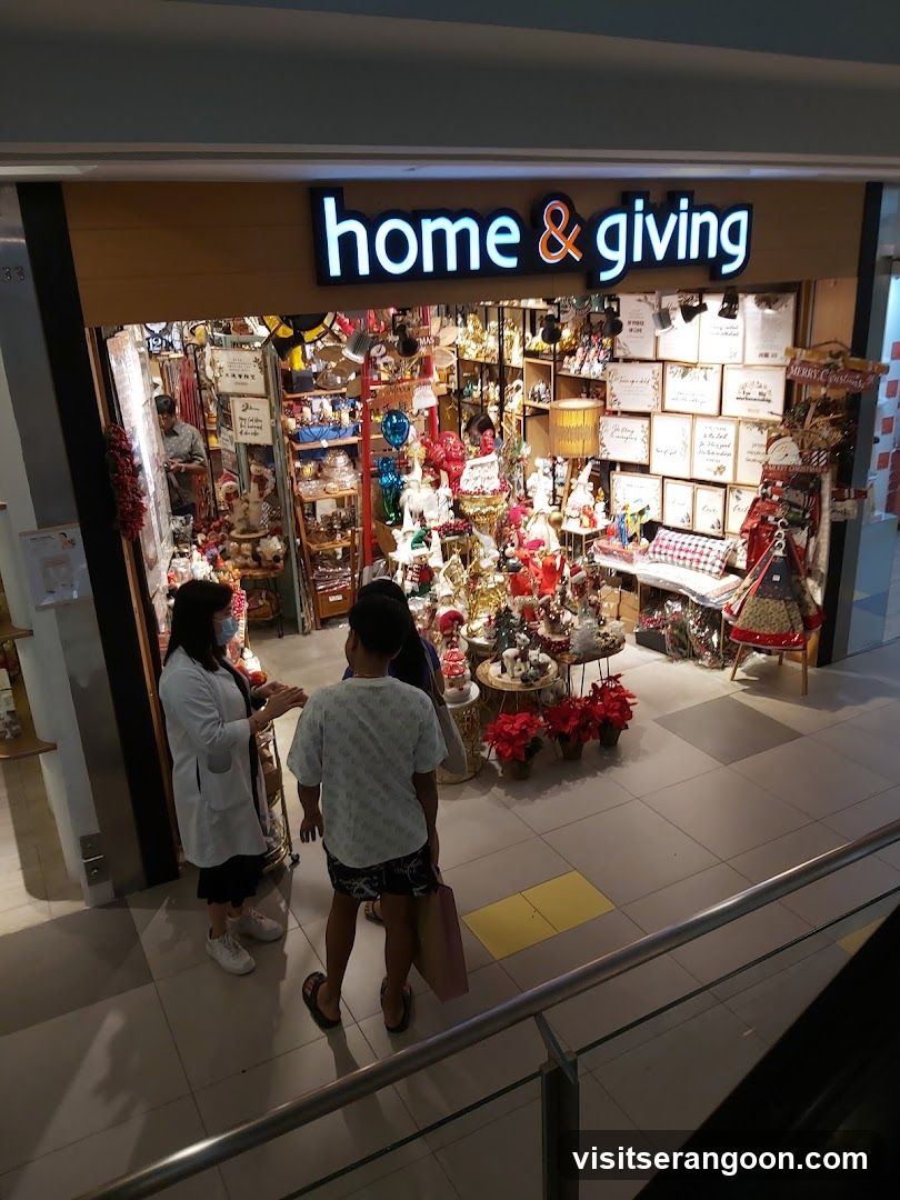 A view from an upper level looking down at the entrance of a retail store named 'home & giving'. The store has a wooden facade with a large, illuminated sign displaying the name in white and orange letters. The entrance is open, revealing a densely packed interior filled with various merchandise, including Christmas decorations, figurines, and home goods. Three people are standing in front of the store on the tiled mall floor, looking inside.