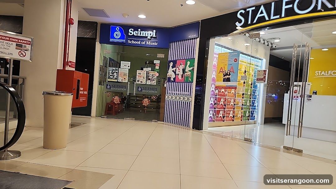 A view of a shopping mall corridor showing two adjacent storefronts. On the left is the Seimpi School of Music, identifiable by its blue signage and glass entrance. On the right is a store named Stalforn, featuring a black facade, glass doors, and a display window filled with colorful posters.