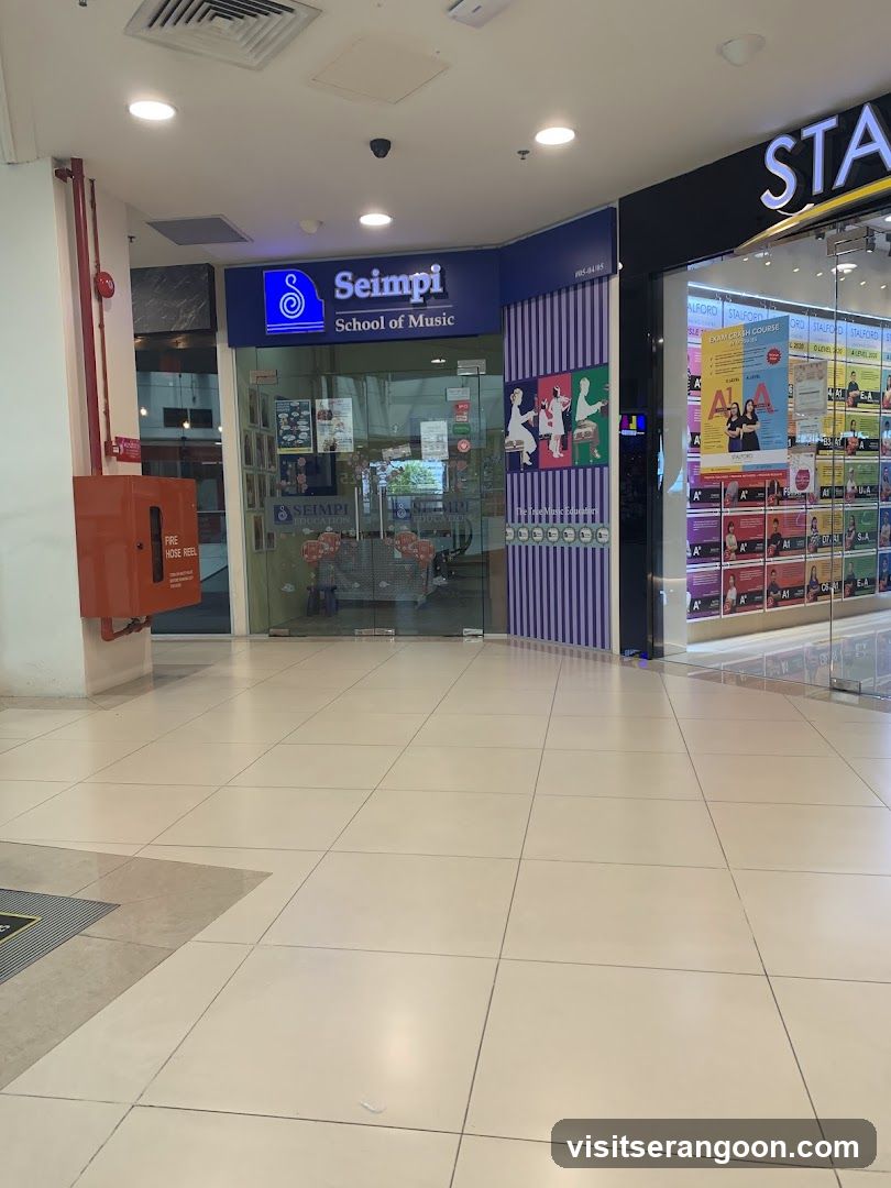 A view of a storefront inside a shopping mall. The central shop is 'Seimpi School of Music', identifiable by its blue sign with a musical note logo. The entrance features glass doors and windows. To the right, a partial view of another store with 'STA' on the sign is visible, displaying colorful posters in the window. To the left, a red fire hose reel box is mounted on a white pillar.