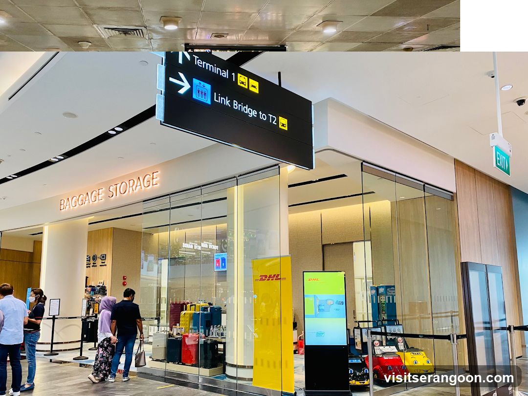 A storefront with a large illuminated sign reading 'BAGGAGE STORAGE' above the entrance. The shop has glass walls and doors, revealing luggage and a DHL counter inside. A large black directional sign hangs from the ceiling pointing to 'Terminal 1' and 'Link Bridge to T2'.