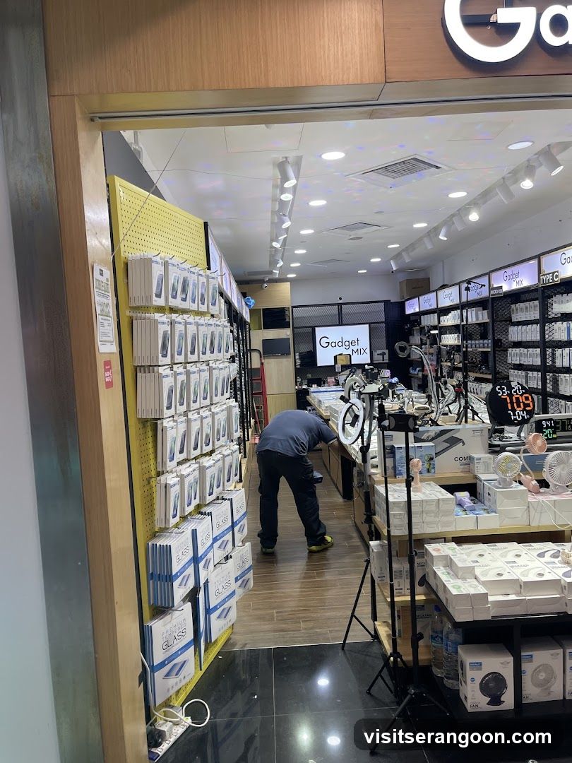 A view into a brightly lit electronics shop named 'Gadget Mix'. The entrance is open, revealing shelves stocked with boxed accessories like phone cases and screen protectors. A staff member is working inside near the counter, and digital price displays are visible on the right.