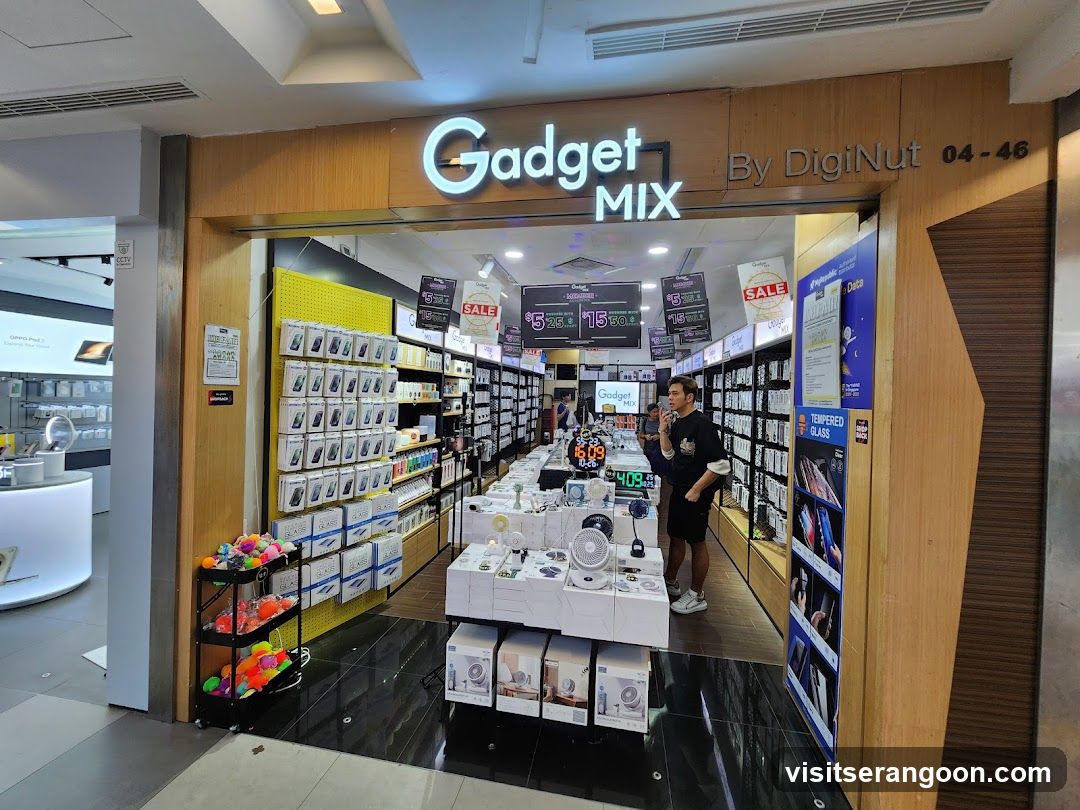 A brightly lit electronics store entrance with a large illuminated sign reading 'Gadget MIX By DigiNut 04-46'. The storefront features wooden paneling and glass doors. Inside, shelves are stocked with boxed gadgets, and promotional signs for sales and prices (e.g., $15, $10) are visible hanging from the ceiling. A man stands near the entrance.