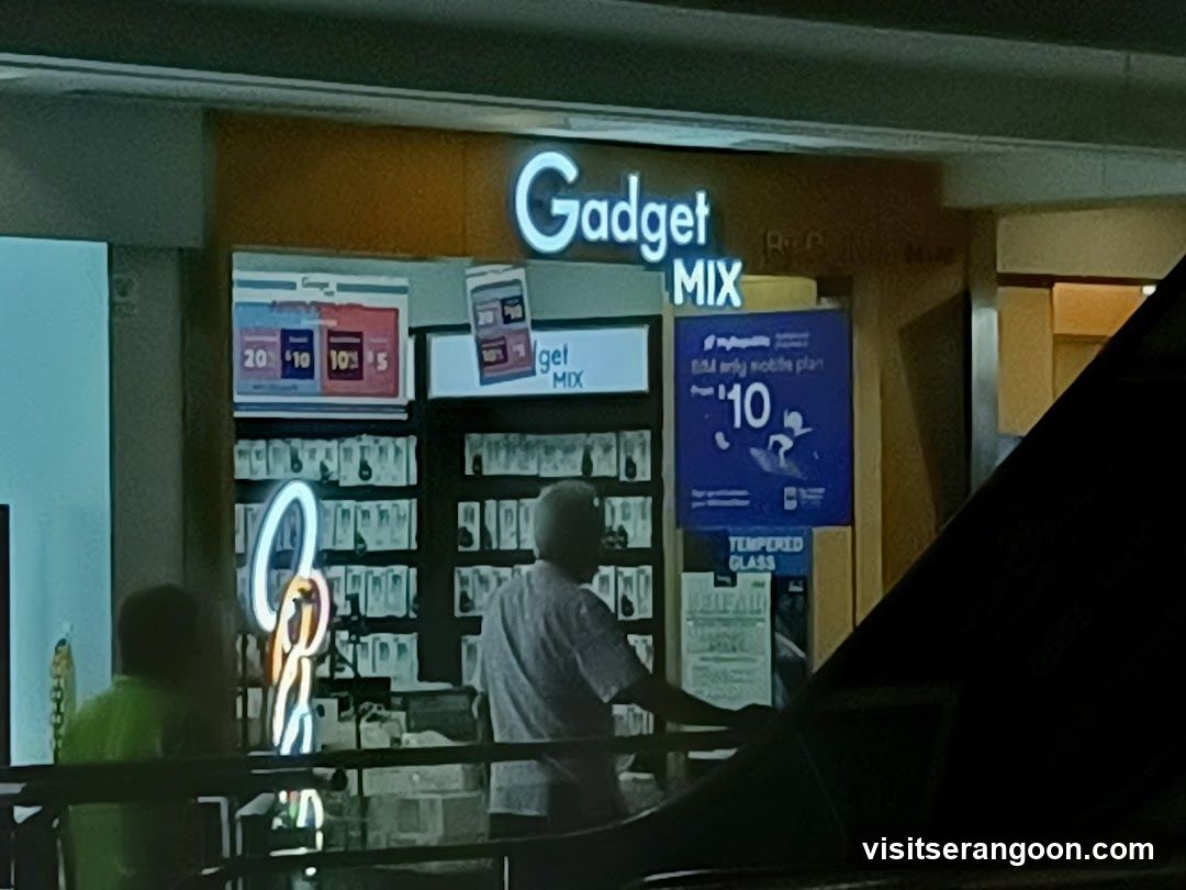 A view of a retail store entrance located inside a building, likely a mall. The store features a prominent illuminated sign reading 'Gadget MIX' in white letters against a yellow background. The storefront displays various posters, including a blue advertisement for a mobile plan starting at $10 and a smaller sign listing prices like $20, $10, and $5. A neon light decoration is visible on the left, and a person is standing inside the store looking at merchandise.