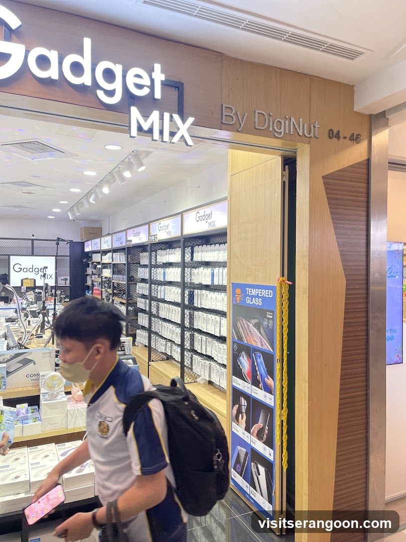A view of a retail store entrance inside a shopping mall. The storefront features a large wooden facade with the illuminated sign 'Gadget MIX' and 'By DigiNut 04-46'. Inside, shelves are stocked with packaged electronics accessories, and a customer wearing a mask and backpack is visible near the entrance.