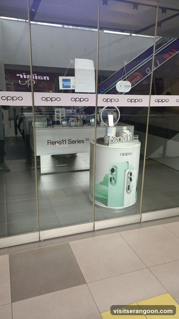 A view through a glass storefront window of an OPPO electronics store. The facade features large glass panels with a white band displaying the 'oppo' logo repeatedly. Inside, a white display counter labeled 'Reno11 Series 5G' and a cylindrical pedestal showcasing a mint green phone are visible. A sign on the glass reads 'Reno11'.