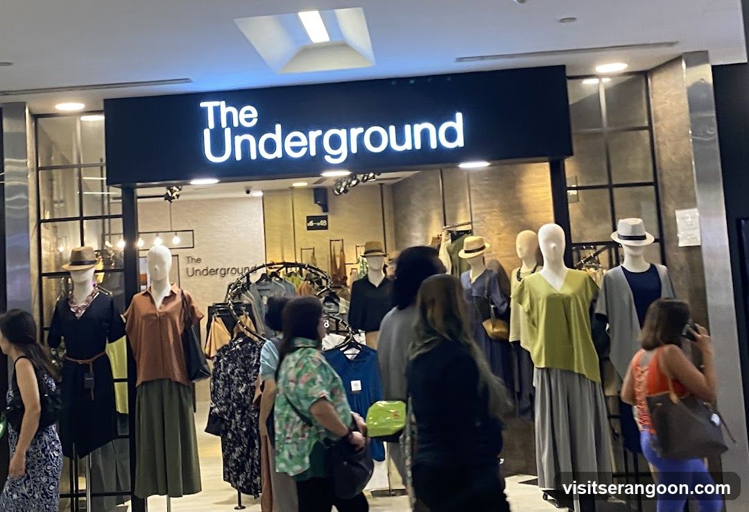 A view of a retail store entrance inside a shopping mall. A large black sign with white illuminated text reads 'The Underground'. The storefront features glass walls and displays several mannequins dressed in clothing, including dresses, skirts, and hats. Several customers are browsing near the entrance.