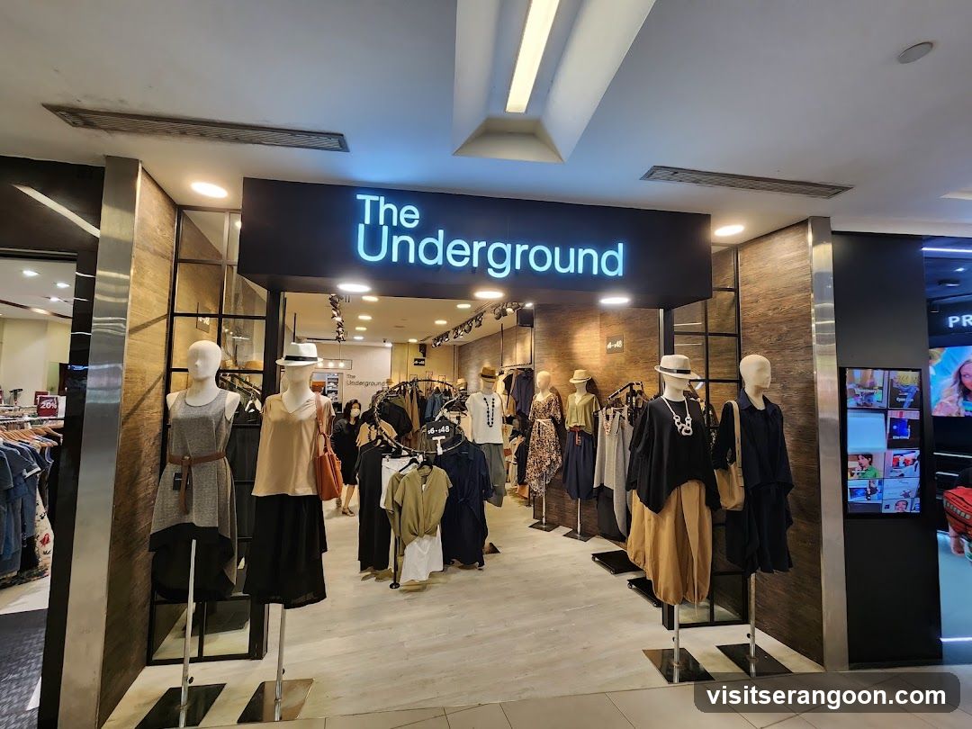A brightly lit retail store entrance featuring a large black sign with the name 'The Underground' in glowing blue letters. The entrance is framed by dark wood paneling and metal pillars. Several mannequins dressed in various outfits, including dresses, skirts, and tops, are displayed on stands at the entrance. Inside, racks of clothing are visible, along with track lighting and a light-colored floor.