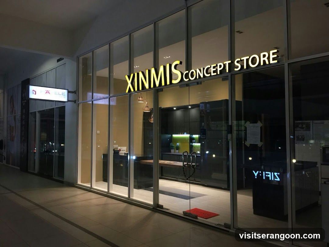 A nighttime photograph of a modern storefront with large glass windows and doors. The business name 'XINMIS CONCEPT STORE' is displayed in large, illuminated yellow letters across the top of the glass facade. Through the glass, a well-lit interior is visible, featuring dark cabinetry, a wooden countertop, and pendant lighting. A red welcome mat sits on the floor inside the entrance. To the left, a portion of an adjacent store with a lit sign is visible.