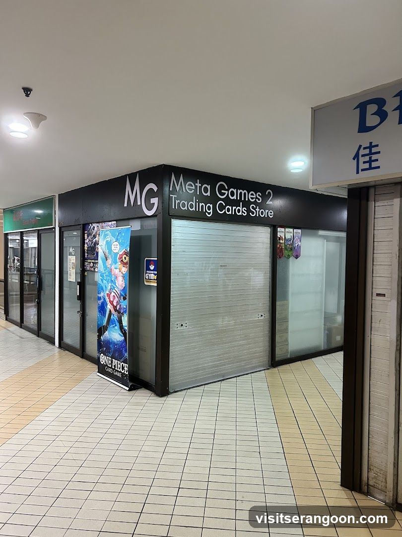 A corner storefront with black cladding and a sign reading 'MG Meta Games 2 Trading Cards Store'. The main entrance is covered by a closed metal shutter. A vertical banner featuring anime characters stands on the tiled floor to the left of the shutter.