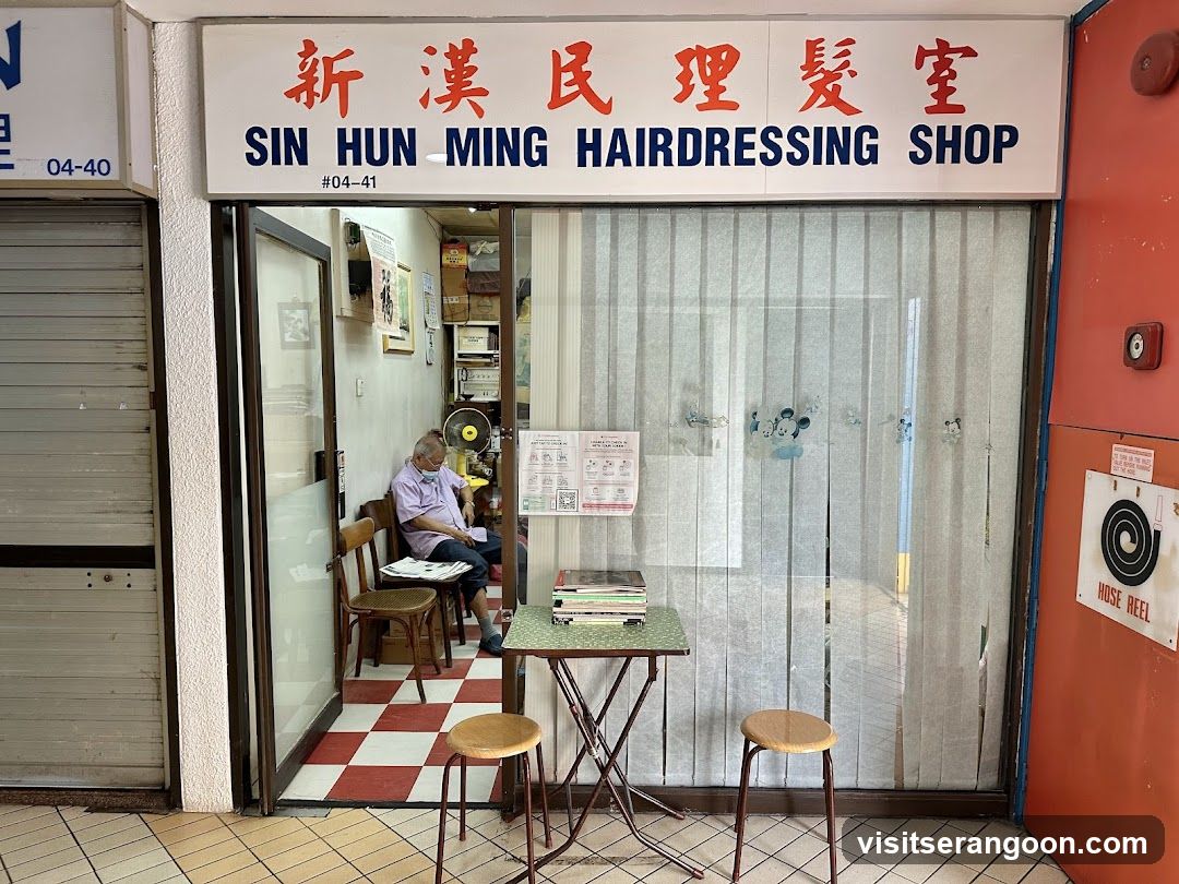A storefront view of a hair salon with a large white sign displaying the name in red Chinese characters and blue English text. The entrance is open, revealing a man sitting inside on a wooden chair. Outside, a small green table and two stools are placed on the tiled pavement. A white curtain covers the window to the right.