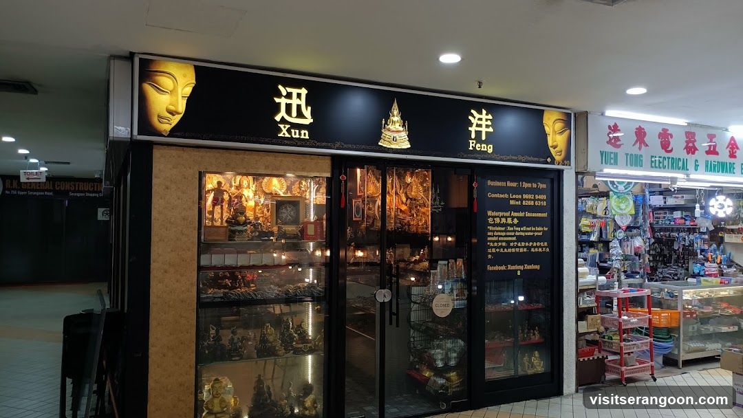 A storefront with a black sign featuring golden Buddha heads and the name 'Xun Feng'. The shop has glass doors and display cases filled with various religious statues and items. A sign lists business hours and contact information. Adjacent to it is another store named 'Yuen Tong Electrical & Hardware'.