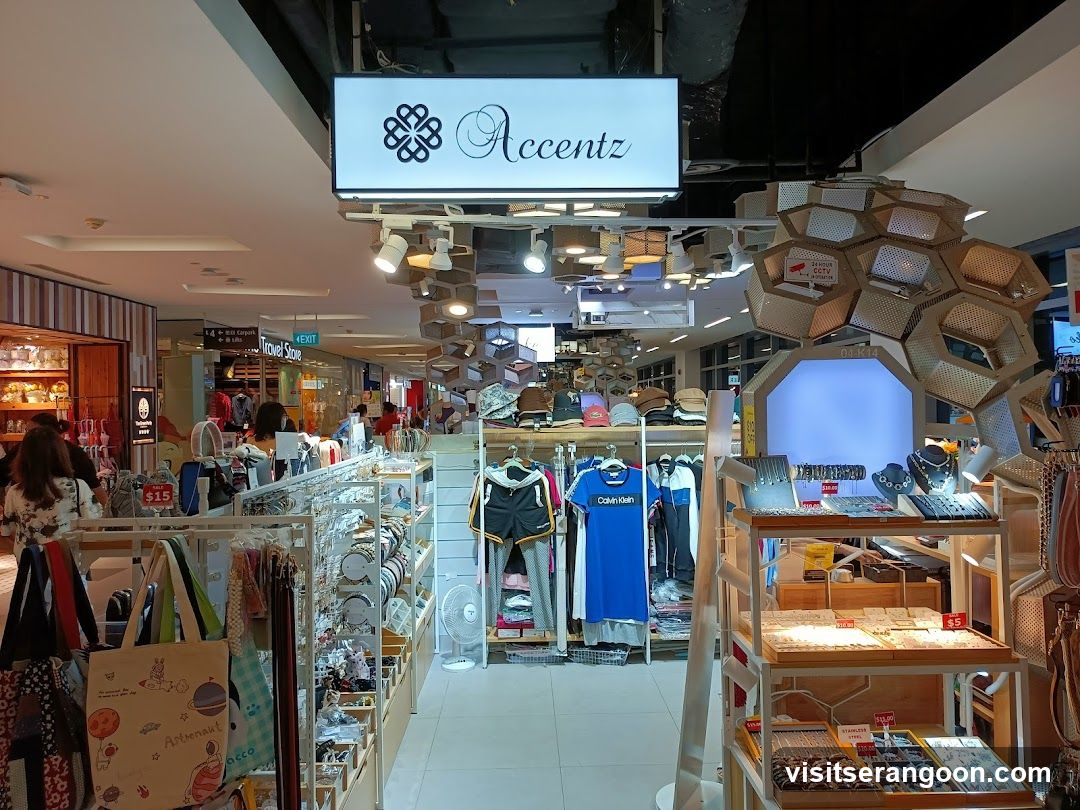A brightly lit retail store entrance inside a shopping mall. A large white sign with the name 'Accentz' and a logo hangs above the entrance. The store displays various merchandise including clothing on racks, bags, and jewelry on wooden shelving units. A distinctive honeycomb-shaped display structure is visible on the right.