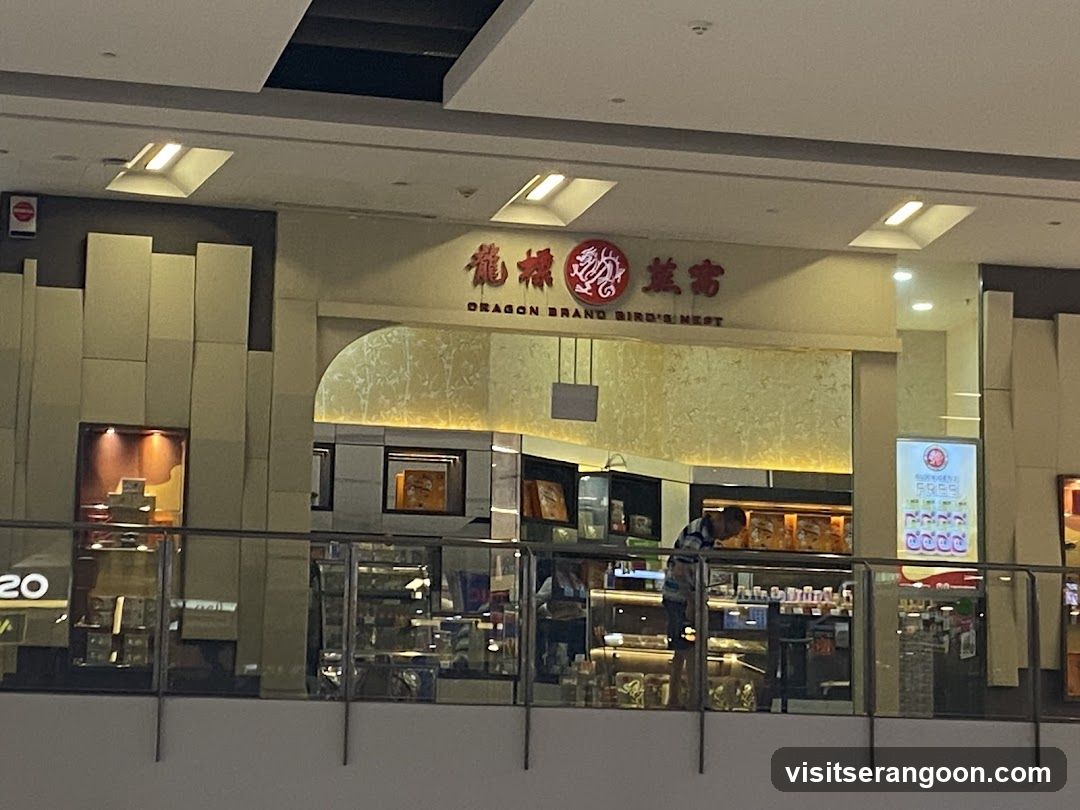 A view of a retail storefront located on an upper level of a shopping mall, viewed through a glass railing. The store features a beige facade with a large arched entrance. The signage above the entrance displays the name '龍標燕窩' in red Chinese characters alongside 'DRAGON BRAND BIRD'S NEST' in English, with a red circular dragon logo in the center. Inside, the store is brightly lit with warm lighting, showcasing glass display counters and shelves stocked with various packaged goods. A staff member is visible behind the counter, and a digital menu board stands to the right of the entrance.