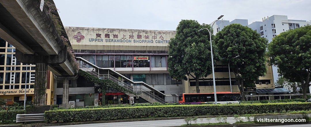 A wide shot of the exterior of the Upper Serangoon Shopping Centre, a multi-story building with concrete architecture. The building name is displayed in large letters on the facade. A prominent external staircase leads to the upper level. A red bus is visible on the street in front of the building, and a concrete overpass structure is on the left side.