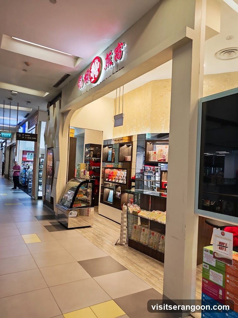 A view of a retail store entrance located in a shopping mall corridor. The storefront features a prominent sign with red illuminated Chinese characters and a circular logo above the entrance. Inside, the store is well-lit with wooden flooring and displays shelves stocked with various packaged goods, likely snacks or health products. A refrigerated display case is visible near the entrance, and a promotional stand with 'BUY 1 GET 1' signage is in the foreground.