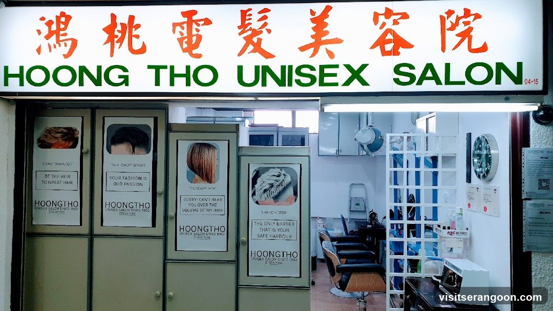 A brightly lit storefront for Hoong Tho Unisex Salon, featuring a large white sign with red Chinese characters and green English text. The entrance is flanked by beige cabinets displaying posters of various hairstyles and salon services. Inside, salon chairs, a white shelving unit, and a wall clock are visible.
