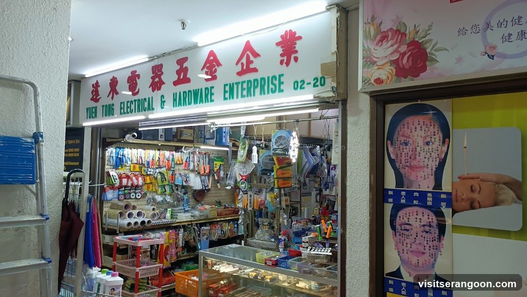A brightly lit storefront for a hardware and electrical shop. The main sign features large red Chinese characters and green English text reading 'YUEN TONG ELECTRICAL & HARDWARE ENTERPRISE 02-20'. The open entrance reveals shelves packed with various goods, including hanging tools, rolls of tape, and items in a glass display case. To the left of the entrance stands a blue step ladder. On the wall to the right, there is a poster featuring a diagram of a human face with acupuncture points.