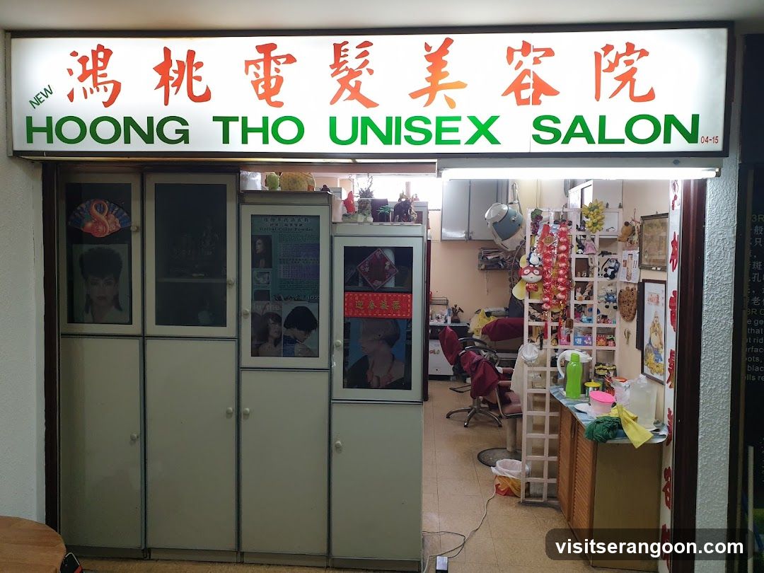 A storefront with a large illuminated sign reading '鴻桃電髮美容院' and 'HOONG THO UNISEX SALON'. The entrance is open, revealing the interior with salon chairs, mirrors, and product displays. To the left of the entrance are tall storage cabinets with framed photos of hairstyles on the doors.